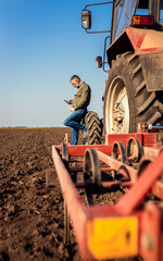 Farmer using smart phone while resting after cultivation of soil leaning on tractor tire.