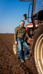 Portrait of satisfied farmer standing next to the tractor in the agricultural field.