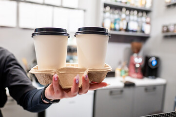 Person holding two takeaway coffee cups in a carrier, with a blurred café interior in the background, symbolizing convenience and on-the-go lifestyle