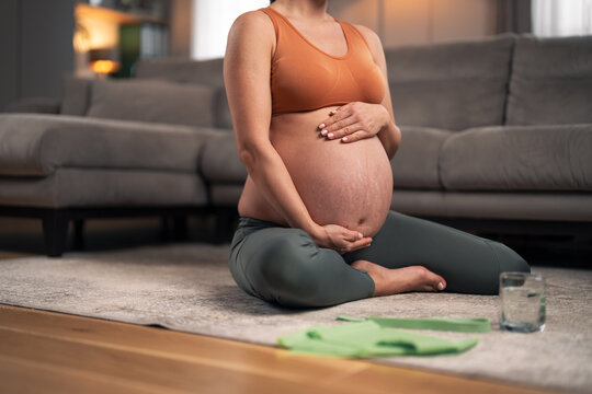 An unrecognizable pregnant woman sitting on the ground in the living room and holding her belly while exercising
