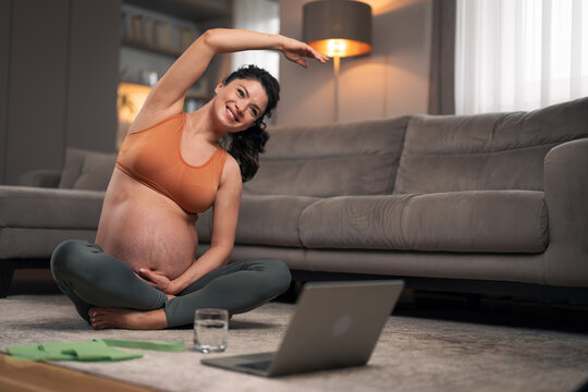 A beautiful pregnant woman doing stretching exercises and following a tutorial on her laptop in the living room