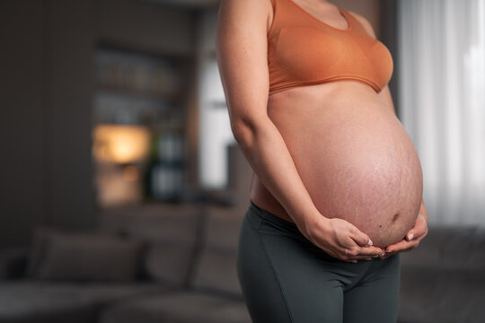 A close-up shot of an unrecognizable pregnant woman holding her belly and standing in the living room