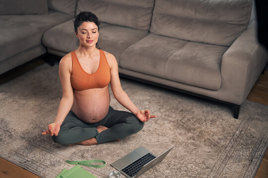 A focused adult pregnant woman following a meditating tutorial on her laptop while sitting on the ground in her living room