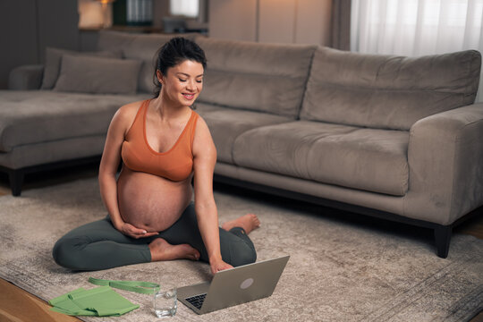 A motivated adult pregnant woman preparing to exercise at home by following a video tutorial on her laptop