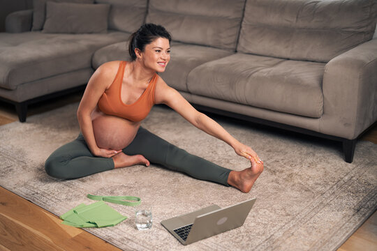 A smiling adult pregnant woman exercising and stretching her body at home