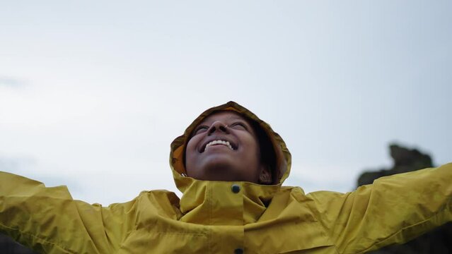 Young black woman standing outdoors in nature under the rain wearing a yellow raincoat, feeling freedom and grateful, raising arms and looking up to the sky with a big toothy smile.