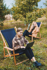 Concentrated woman in the garden working on a laptop while sitting on a chair among the trees. A woman works remotely from the garden, thinking about a new business or creative idea.