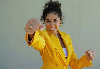 A cheerful and self-assured Latina businesswoman confidently poses in a vibrant yellow suit, extending one arm forward in a dynamic gesture towards the camera.