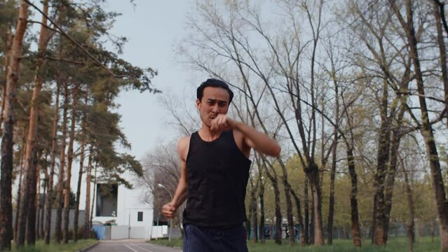 Young asian male athlete running and making punches in front of himself at the same time during workout on an alley in an autumn city park. Concept of modern outdoor sports training in a casual urban