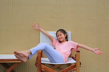 Smiling Asian girl child open hands outstretched and relaxing on wood chair against cement wall background. - 793793752