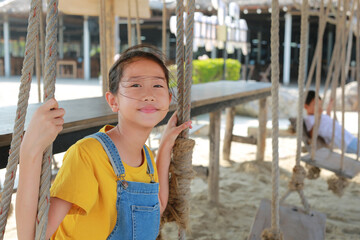 Close up of Asian girl in dungarees jean with wind blowing hair on face sitting on swing at cafe near coast.