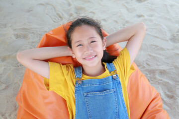Close-up smiling of Asian young girl in dungarees jean looking camera while relax on orange sofa bed beach on sand at summer holiday. - 793796534