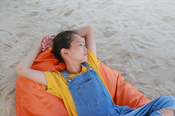 Portrait of Asian young girl in dungarees jean looking beside and relax on orange sofa bed beach on sand at summer holiday.