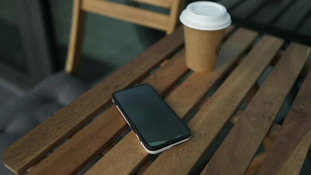 Paper cup of coffee and mobile phone on a brown wooden table, close-up outdoors. Eco paper glass of coffee and a mobile phone lie on a brown wooden table surface. High quality FullHD footage