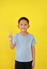Portrait of Asian boy child show victory sign fingers and looking straight while standing isolated on yellow background.