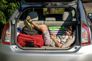 Cute little boy laying on the back of the bags and baggage in the car trunk ready to go on vacation with happy expression. Kid resting playing on smartphone - obrazy, fototapety, plakaty
