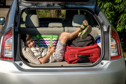 Cute little boy laying on the back of the bags and baggage in the car trunk ready to go on vacation with happy expression. Kid resting playing on smartphone