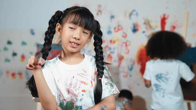 Creative cute girl pose at camera while diverse children painting behind. Young beautiful child looking at camera while standing at stained room and holding paintbrush. Creative activity. Erudition.