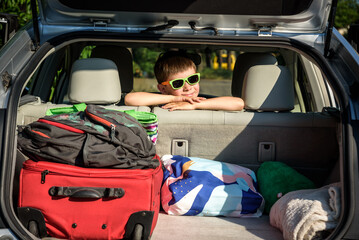 Adorable kid boy wearing sunglasses sitting in car trunk. Portrait of Happy child with open car boot while waiting for parent get ready for vocation. Family trip traveling by car concept