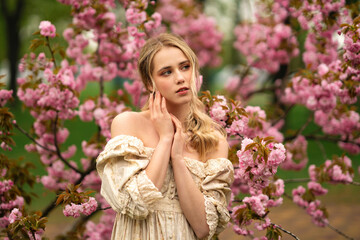 Pretty young blonde girl  in vintage lace dress  standing in spring park near pink blossom flowers....
