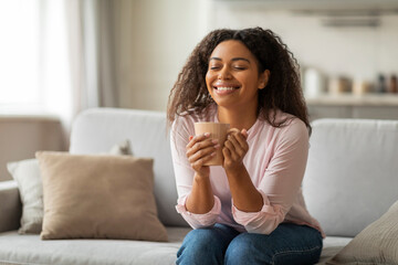 Positive young black woman sitting on couch, drinking coffee