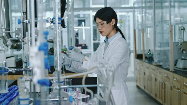 Medium footage of Chinese female scientist in white coat, goggles, gloves adding drops of reagent to chemical solution in vial, holding it up, checking for reaction or residue, while conducting tests