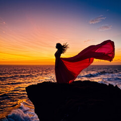 A woman in a red dress stands on a rocky cliff overlooking the ocean. The sky is a beautiful mix of orange and pink hues, creating a serene and peaceful atmosphere