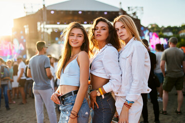 Three girls enjoy summer music festival on sandy beach at sunset. Friends pose for photo, outdoor...
