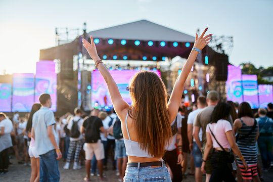 Back view of girl with raised arms, enjoying live music at sunset beach festival. Friends dance, celebrate in crowd, summer vibe at coastal event. Teens in casual wear, fun at sunny seaside gig.
