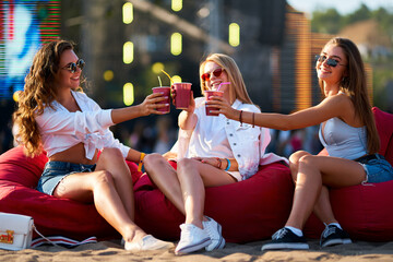 Group of happy young females sit on red beanbags at sunny seaside music festival, cheers with drinks, chill vibes. Three carefree friends in summer outfits, sunglasses clink cups, relax on coast.