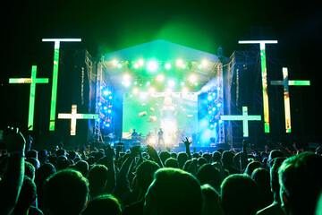 Crowd celebrates at night religious music festival with glowing stage, vibrant green lights, neon crosses. Joyful audience raises hands, enjoys live gospel performance, outdoor worship concert.