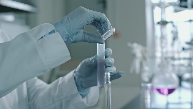 Close-up shot of hands of anonymous female laboratory employee in gloves adding pink chemical solution from tube to clear liquid in vial while performing diagnostic test on biochemical probe