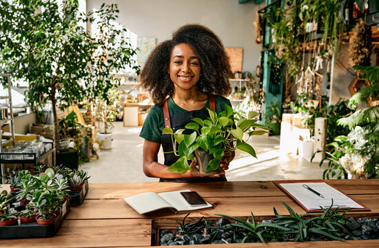 Work in a plant shop. Beautiful african american woman florist gardener holding a flower pot and smiling while standing in a beautiful green space among flower pots, plants and decor.