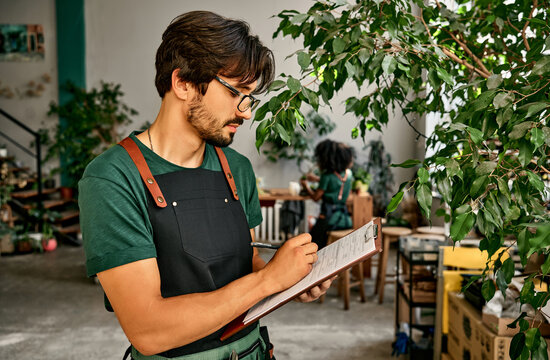 Plant nursery. Side view of bearded man in uniform and glasses writing on clipboard while standing in spacious shop with green plants. Caucasian florist making notes during inventory process at work.