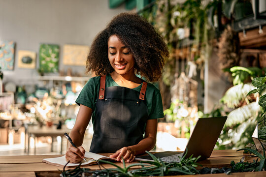 Technology for business. Beautiful african woman using wireless laptop and writing on clipboard at modern flower boutique. Positive female owner analyzing stock of plants at favorite small business.