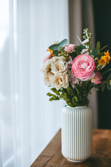 A lively bouquet of flowers, adorns a white, ribbed vase atop a wooden surface, set near the window