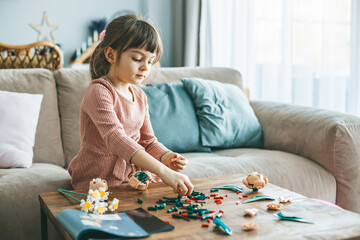 A cute little girl is immersed in play, using small colorful construction pieces on a wooden table to create flowers