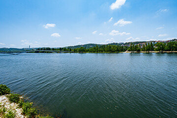 A view of the Mondego River in Coimbra, Portugal, under a clear sky, with trees and a pedestrian...