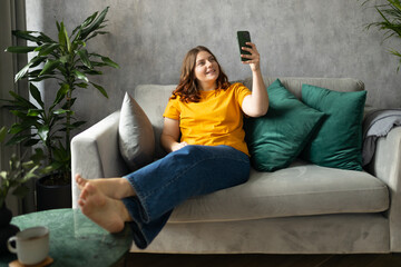 Hi. Brunette taking a selfie photo using her mobile phone at home. 30s woman in yellow t-shirt recording self video, talking on online call and relaxing on a sofa. Full body