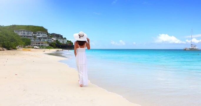 A elegant woman in a white summer dress walks along the beautiful Darkwood beach at the Caribbean island of Antigua
