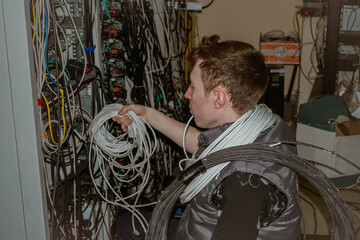 A technician with a bundle of cables around his neck works in a server room. A man lays a cable at a telephone exchange