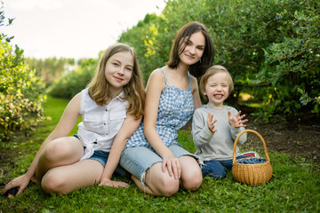 Cute teenage sisters and their little brother picking fresh berries on organic blueberry farm on warm and sunny summer day. Fresh healthy organic food for kids. - 793868784
