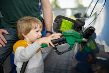 Cute little blond boy holding pump nozzle. Small funny kid helping father to fuel the car at a gas station. - 793870140