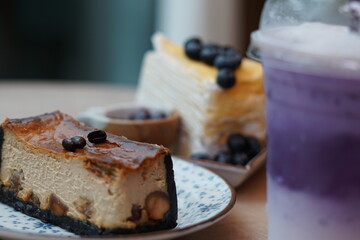 A piece of espresso macadamia cheese cake topped with coffee beans on the plate with blur of blueberry crepe cake and sweet potato milk tea.