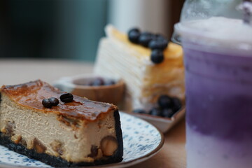 A piece of espresso macadamia cheese cake topped with coffee beans on the plate with blur of blueberry crepe cake and sweet potato milk tea.