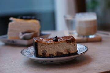 A piece of espresso macadamia cheese cake topped with coffee beans on the plate with blur of blueberry crepe cake and dirty latte coffee.