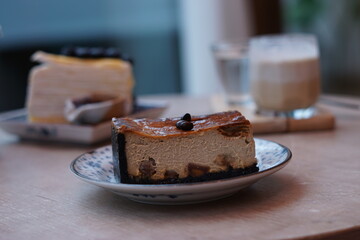 A piece of espresso macadamia cheese cake topped with coffee beans on the plate with blur of blueberry crepe cake and dirty latte coffee.