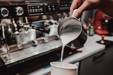 Hand pouring milk from a steel pitcher into a coffee cup, with an espresso machine in the background, capturing the art of coffee making