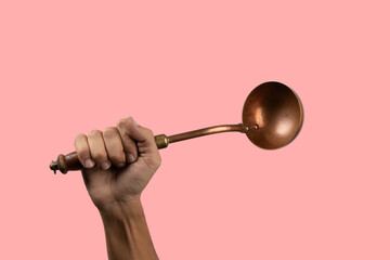 Black male hand holding a brass vintage kitchen ladle isolated on pink background