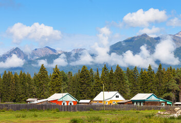 Village wooden houses against backdrop of spruce forest and Eastern Sayan Mountains with low clouds on Tunka foothill valley on sunny summer day. Scenic rustic landscape. Siberia. Buryatia. Arshan